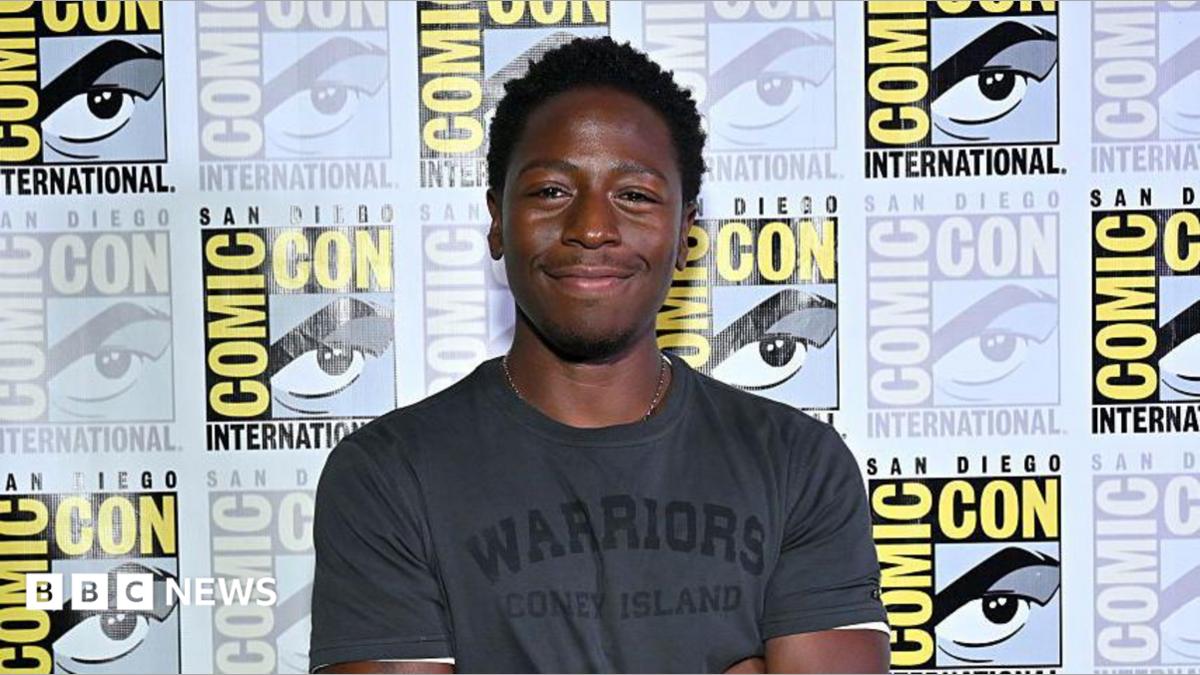 David Jonsson, the new Black Panther, stands in front of a Comic-Con International hoarding featuring a repeating pattern of