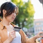 A woman uses a cash machine on the street on a sunny spring day. She holds a credit card in her hand and is pressing buttons