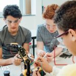 Three students wearing safety goggles work together on a robotic arm in a classroom or laboratory, connecting wires and