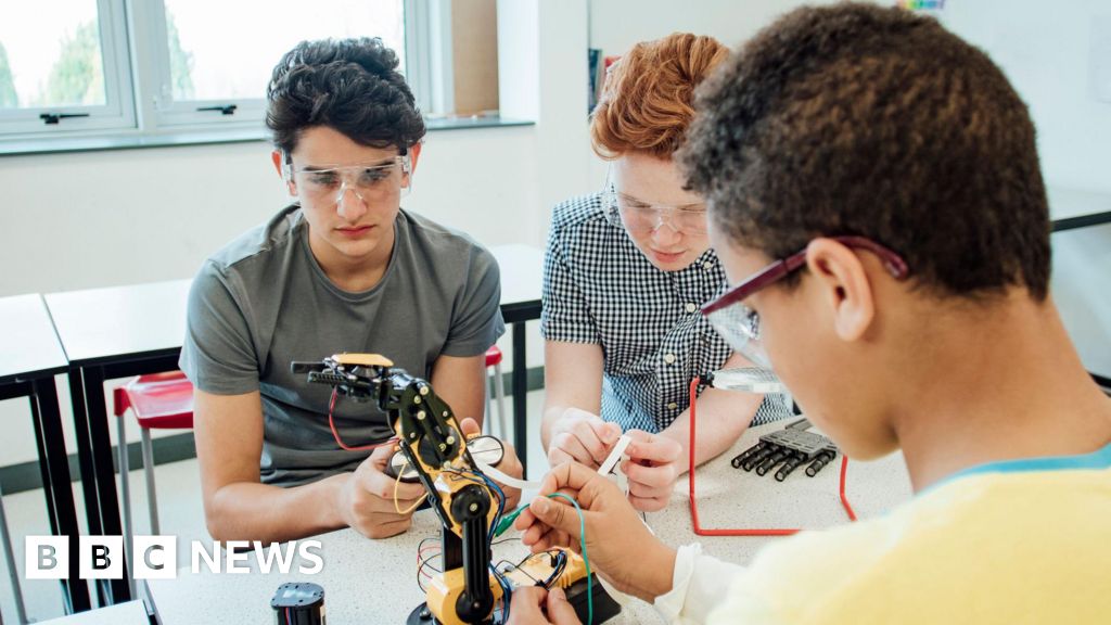 Three students wearing safety goggles work together on a robotic arm in a classroom or laboratory, connecting wires and