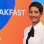 Naga Munchetty posing and smiling in front of the BBC Breakfast studio and logo