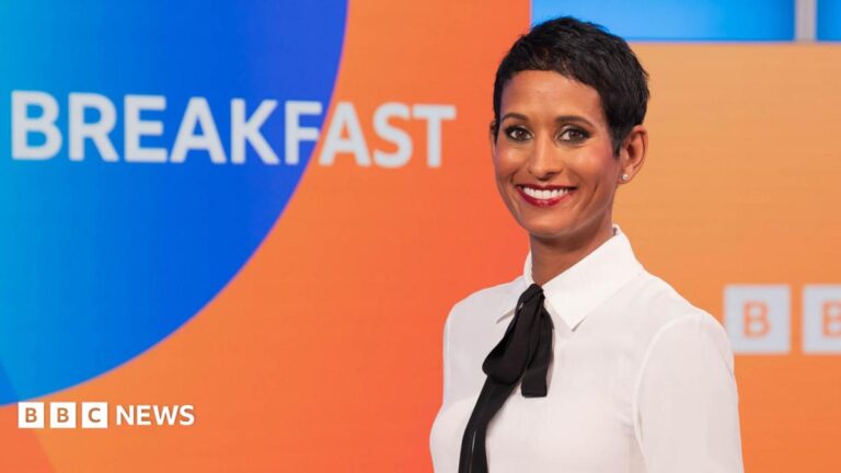 Naga Munchetty posing and smiling in front of the BBC Breakfast studio and logo