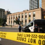 An image showing two Toronto Police officers standing behind yellow caution tape across a three-lane road. Behind them is a