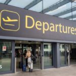 The departures entrance at Gatwick Airport with large yellow "Departures" signage above glass doors. Travellers with luggage