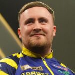 Luke Littler, pictured in March 2026, wearing a blue and yellow jacket during a darts tournament