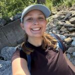 Megan Szostak stands in a rocky stream, taking a selfie and smiling at the camera. She wears a black t-shirt, blue backpack