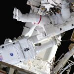 Two astronauts in white bulky spacesuits work outside a large space station set against the blackness of space