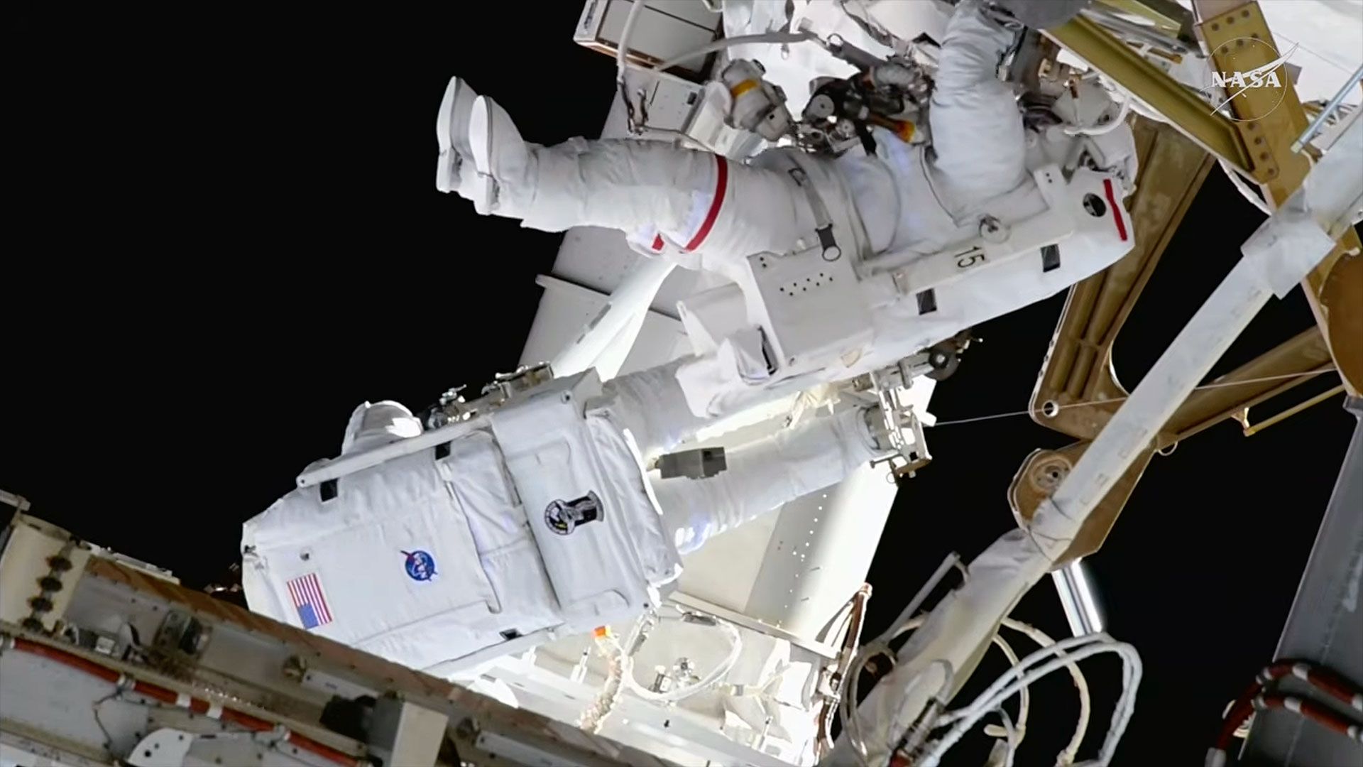 Two astronauts in white bulky spacesuits work outside a large space station set against the blackness of space