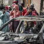 A crowd of people look on close to a burned out car