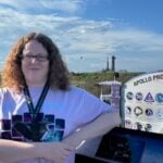 Rebekah Hounsell stands next to an informational placard that reads "Apollo Program," with the SpaceX Falcon Heavy rocket