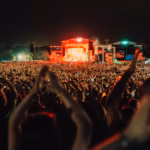 A large crowd at an outdoor festival at night in front of a stage with orange lighting