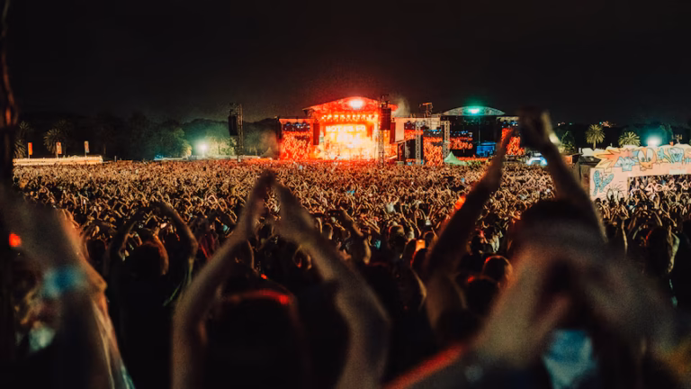 A large crowd at an outdoor festival at night in front of a stage with orange lighting