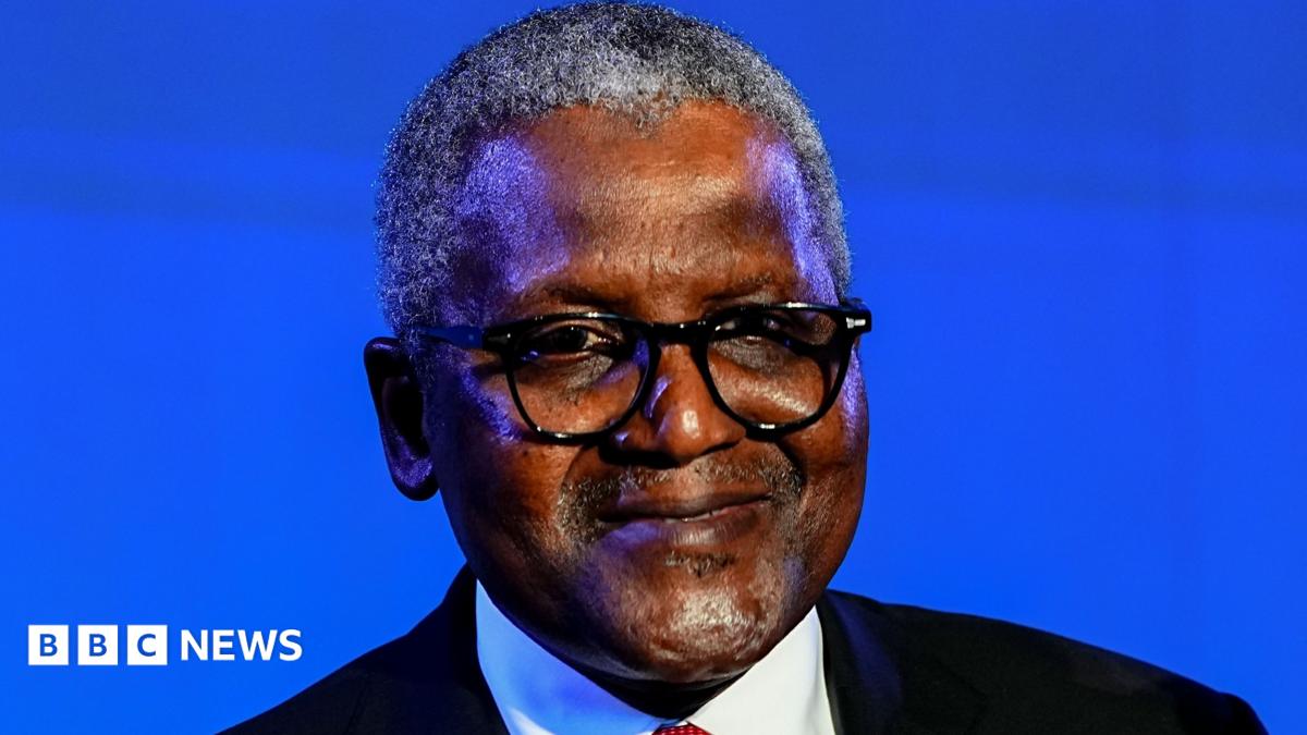 A man with grey hair, wearing a black suit, white shirt, and red tie, smiling into the camera