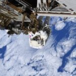 a person in a bulky white spacesuit clings to metal bars attached to a large structure, while white clouds in a blue sky can
