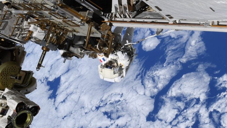 a person in a bulky white spacesuit clings to metal bars attached to a large structure, while white clouds in a blue sky can