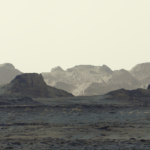 A stark, grayish-tan Martian landscape features jagged rock formations and rolling mountain ridges beneath a pale, hazy sky