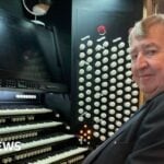 Prof Ian Tracey with short grey hair and wearing a black suit sits at the grand organ inside Liverpool Cathedral, with