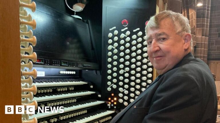Prof Ian Tracey with short grey hair and wearing a black suit sits at the grand organ inside Liverpool Cathedral, with