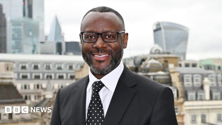 A smiling man in suit and tie standing in front of the City of London skyline