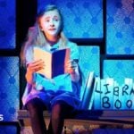 The 2011 child actor playing Matilda sits on a wooden shelf with library book props next to her on stage. She is holding a