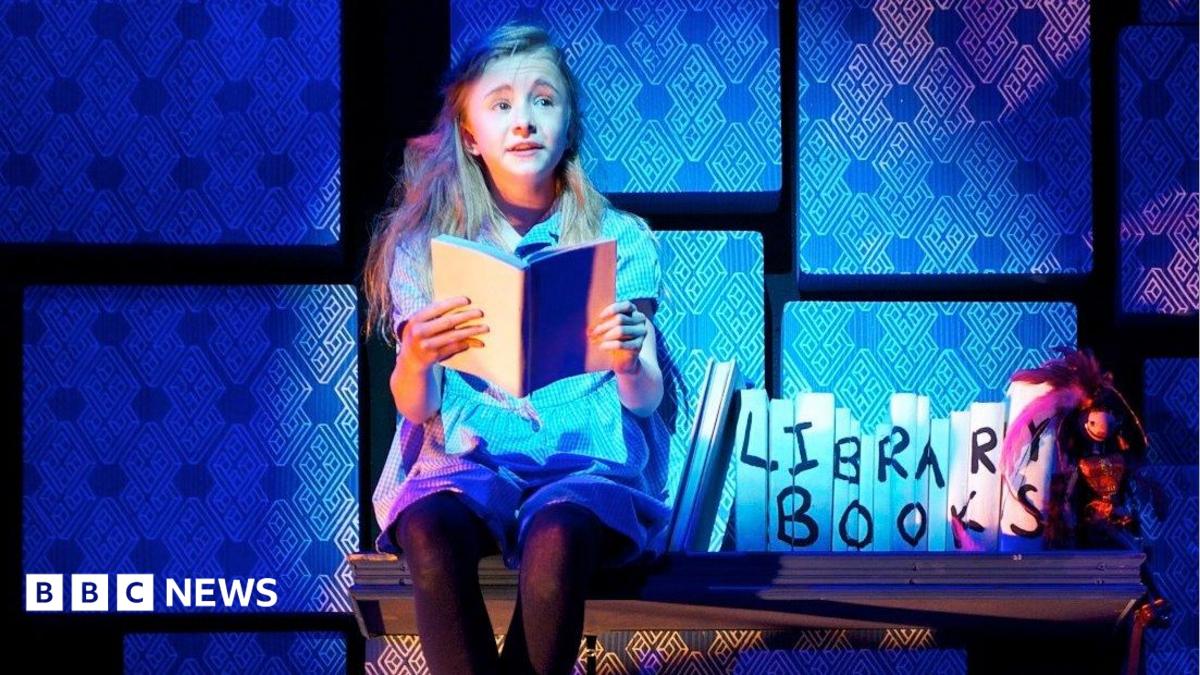 The 2011 child actor playing Matilda sits on a wooden shelf with library book props next to her on stage. She is holding a