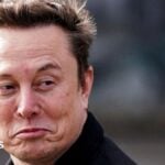 Elon Musk smiles at a camera while appearing to walk by. He is wearing a black coat and has short brown hair