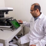 A photo of a researchers in a lab sitting near a machine with a touchscreen monitor