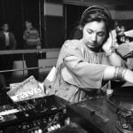 A black and white photo of a young woman spinning a record on DJ decks. She is wearing traditional South Asian clothing as