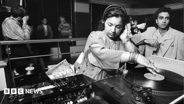 A black and white photo of a young woman spinning a record on DJ decks. She is wearing traditional South Asian clothing as