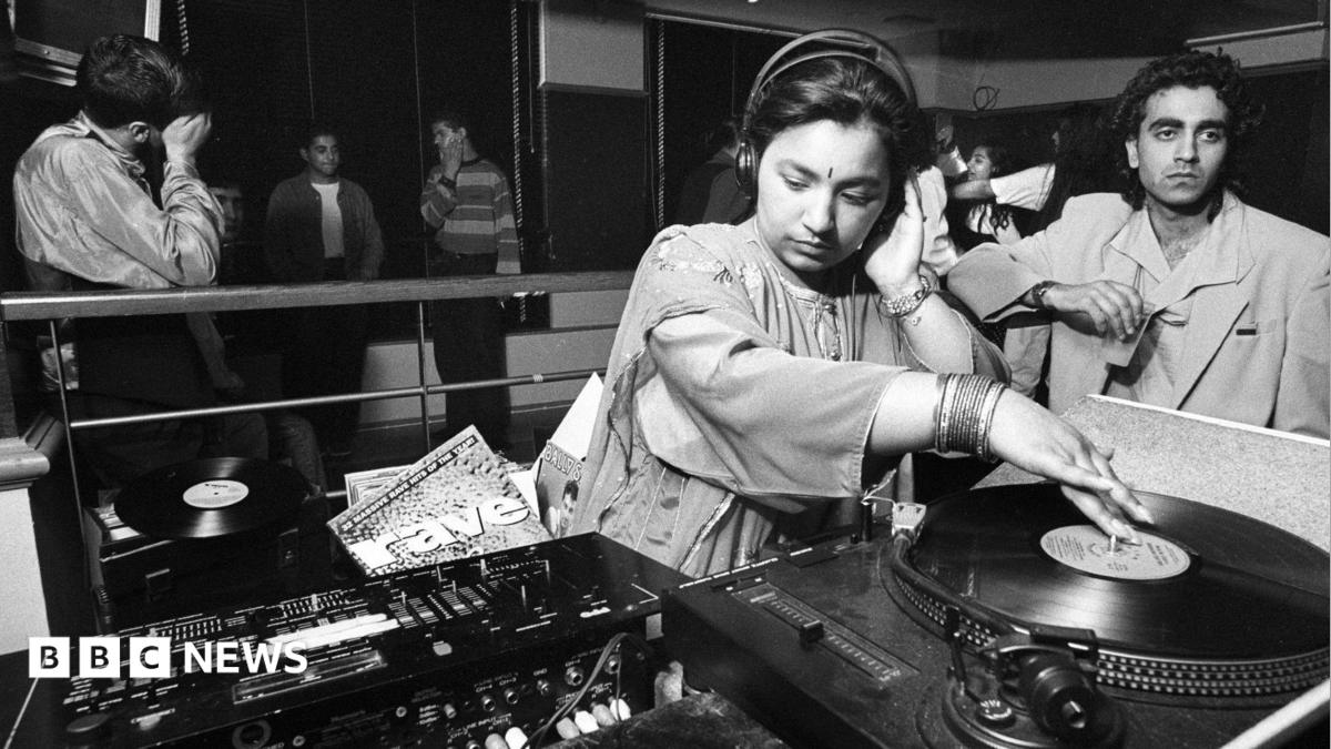 A black and white photo of a young woman spinning a record on DJ decks. She is wearing traditional South Asian clothing as