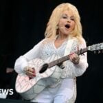 Dolly Parton is holding an acoustic guitar and wearing a white outfit while singing on stage
