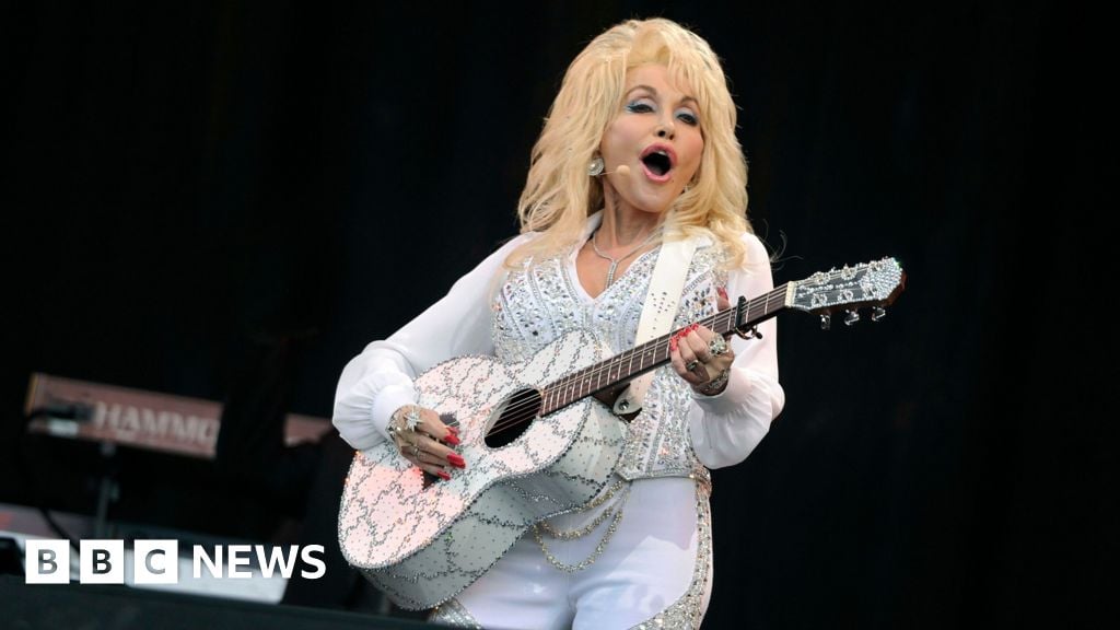 Dolly Parton is holding an acoustic guitar and wearing a white outfit while singing on stage