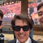 Two men dressed formally pose for a photo with actor Tom Cruise who wears a tuxedo and black sunglasses, at a film preview