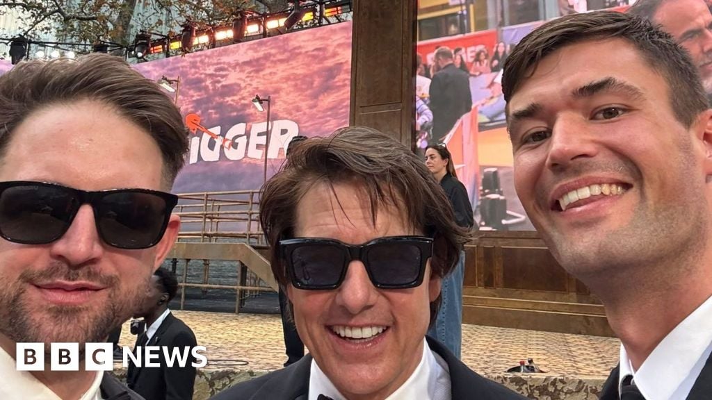 Two men dressed formally pose for a photo with actor Tom Cruise who wears a tuxedo and black sunglasses, at a film preview