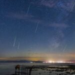 multiple meteors streak through a star studded sky with the milky way off to the right. there are lights shining over the