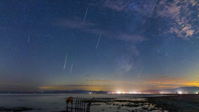 multiple meteors streak through a star studded sky with the milky way off to the right. there are lights shining over the